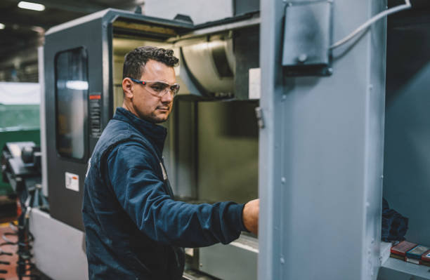 Portrait of apprentice engineering worker young man working, examining and operating CNC plastic injection molding machinery in factory warehouse after studied manufacturing apprenticeship program certifies XXXL